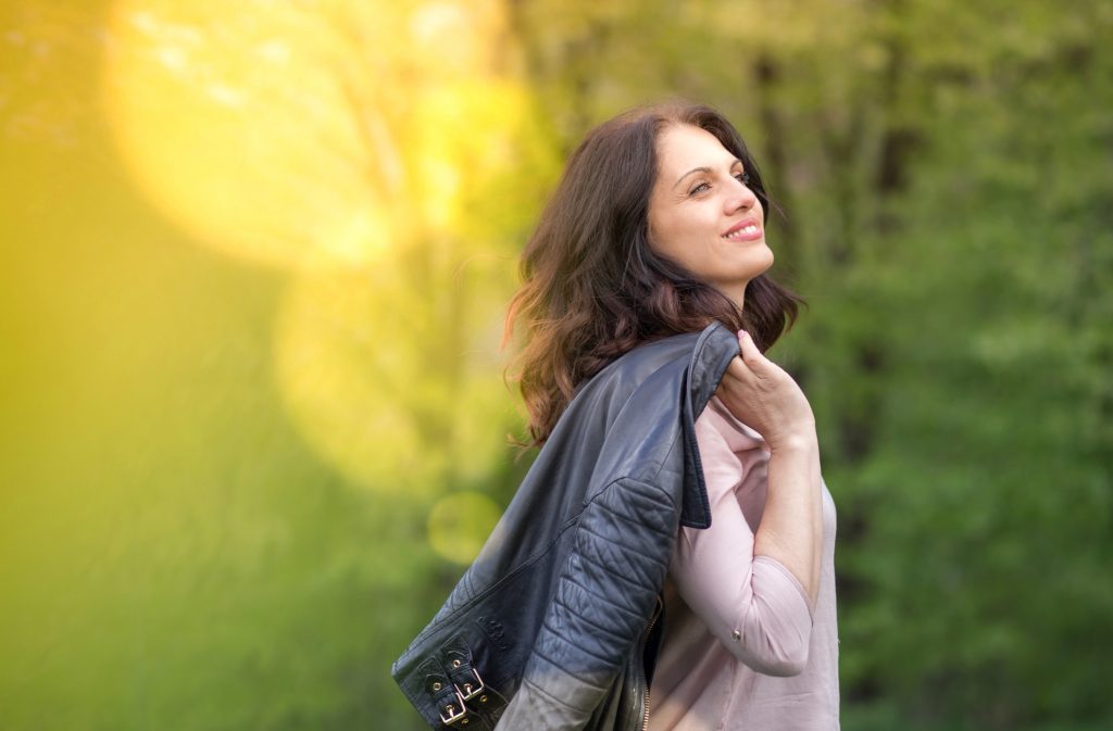 Beautiful, powerful middle-age woman in black leather jacket smiling, holds her jacket over the shoulder. Springtime, outdoors